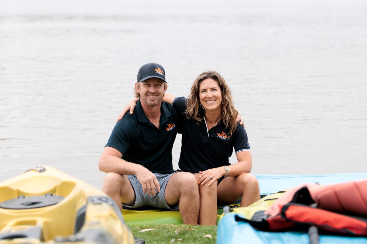 Two people sitting on kayaks by a lake, smiling, wearing matching shirts and caps.