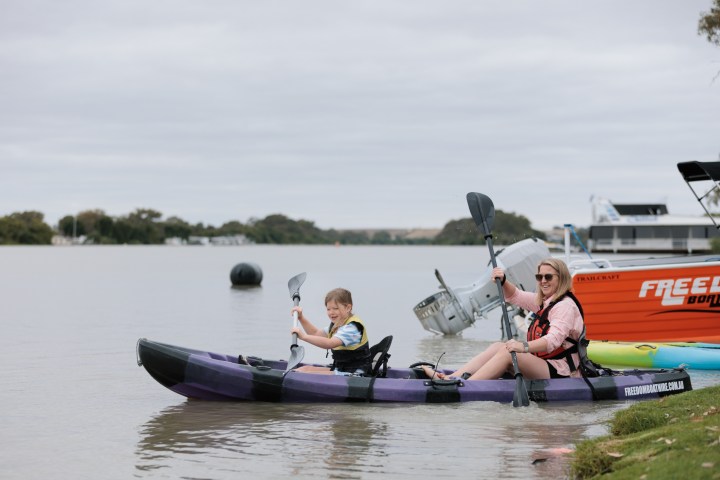 Woman and child kayaking on a calm river near boats and trees.