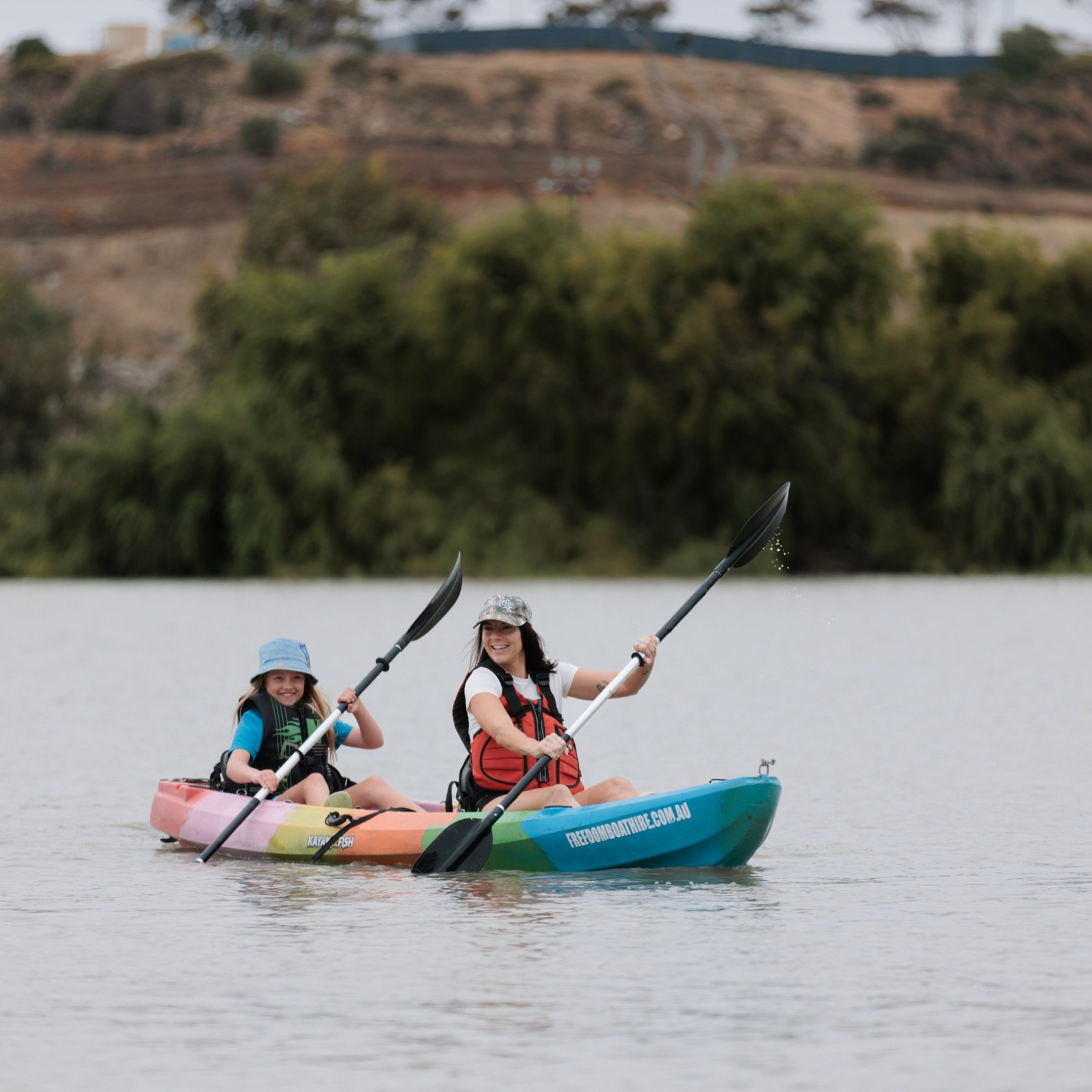 Two people kayaking on a calm lake with trees in the background.