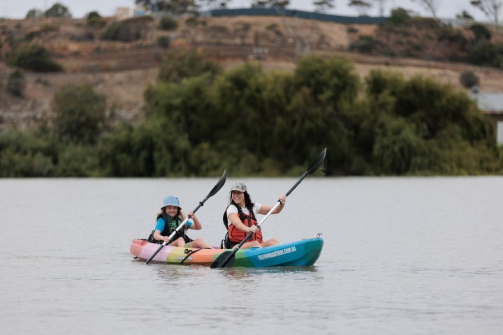 Two people kayaking on a calm lake with trees in the background.