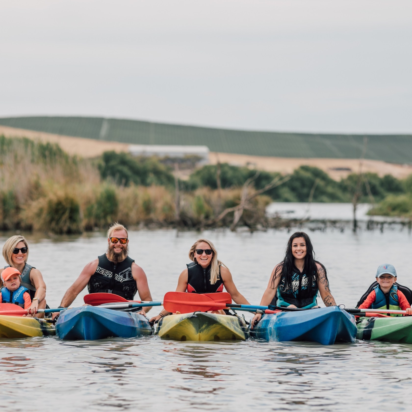 Five people kayaking on a river in colorful kayaks, surrounded by greenery.