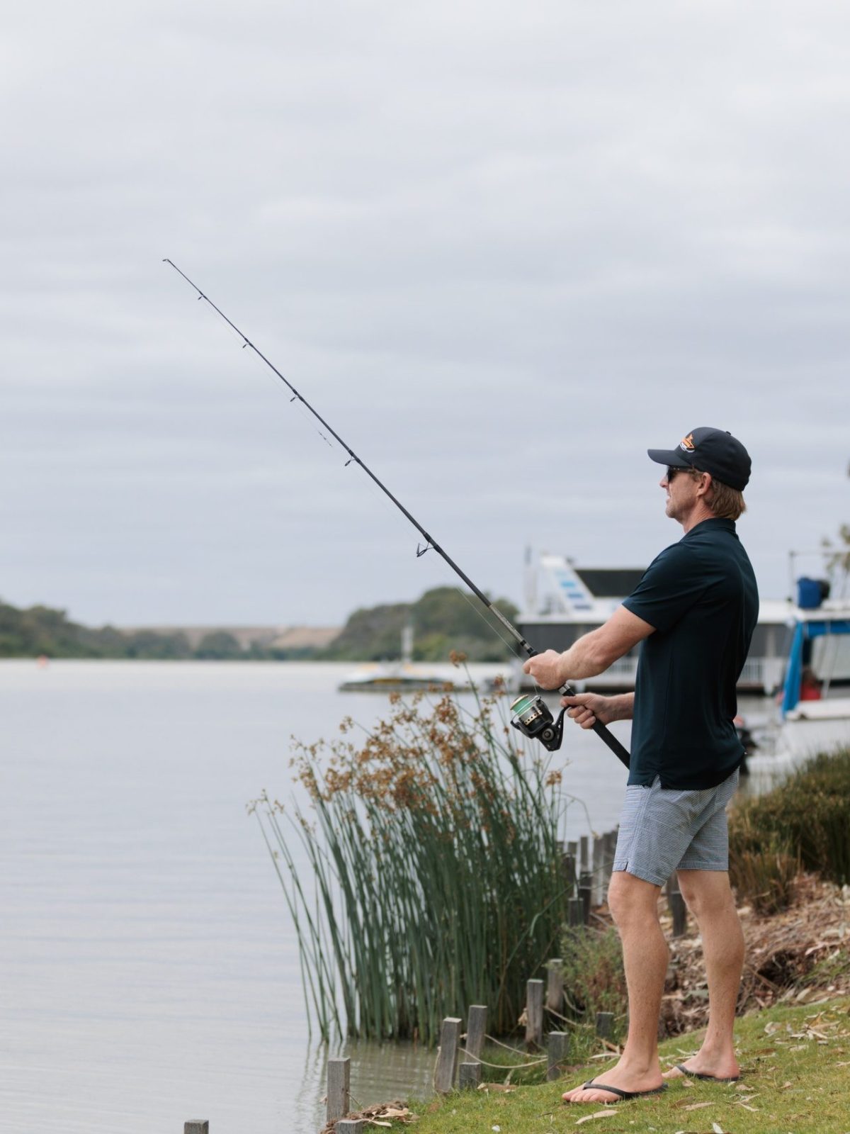 Man fishing by a lake, wearing casual clothes and a cap, boats in the background.