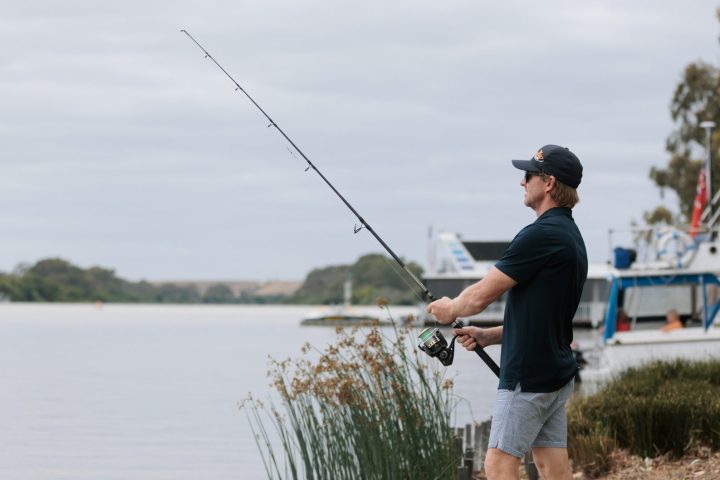 Man fishing by a lake, wearing casual clothes and a cap, boats in the background.