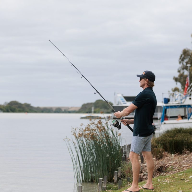 Man fishing by a lake, wearing casual clothes and a cap, boats in the background.