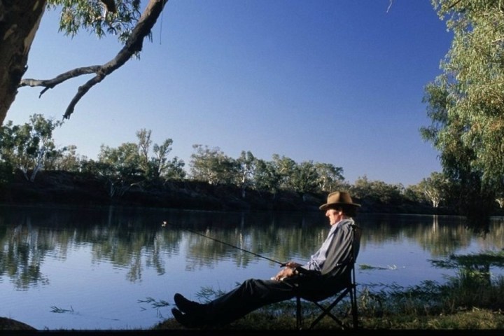 Person fishing by a calm river under a clear blue sky.