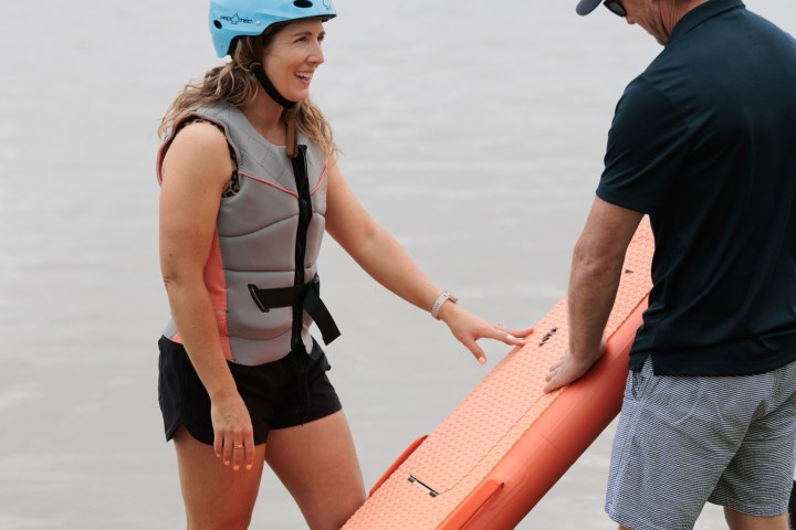 Two people holding an orange paddleboard by a lake.