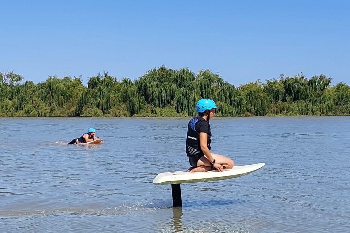 Person kneeling on a hydrofoil board on a lake, wearing a helmet; another person swims nearby.