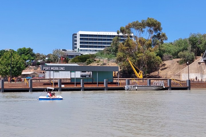 People in paddle boat on river near port building and a bridge.