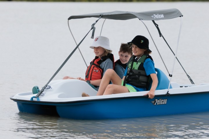 Three children in life jackets ride a small pedal boat on calm water.
