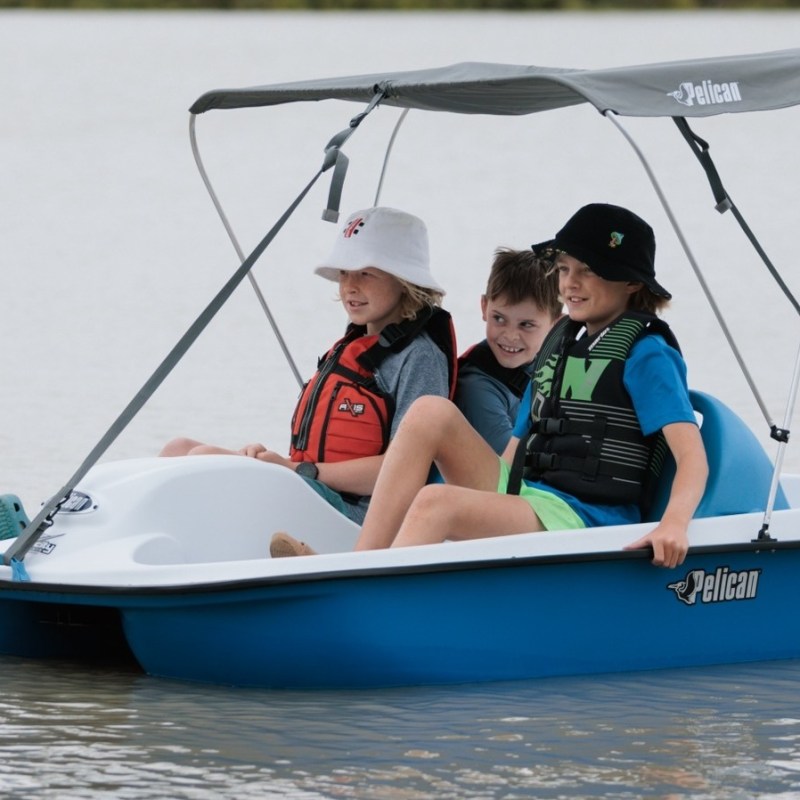 Three children in life jackets ride a small pedal boat on calm water.