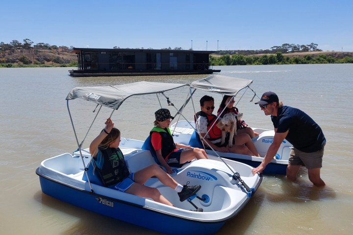 Four people and a dog in paddle boats on a lake, with a houseboat in the background.