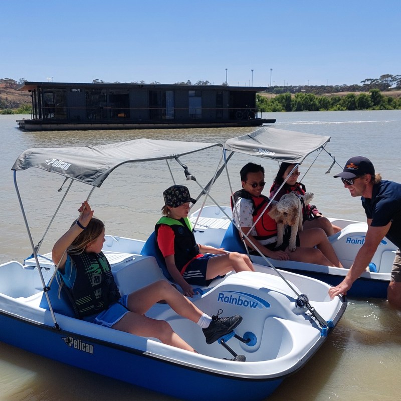 Four people and a dog in paddle boats on a lake, with a houseboat in the background.