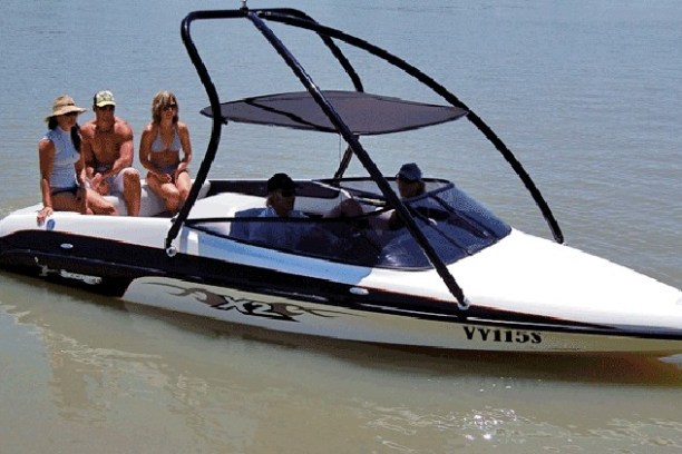 Sleek speedboat with canopy, three people in swimsuits sitting on back, two in front, on calm water.