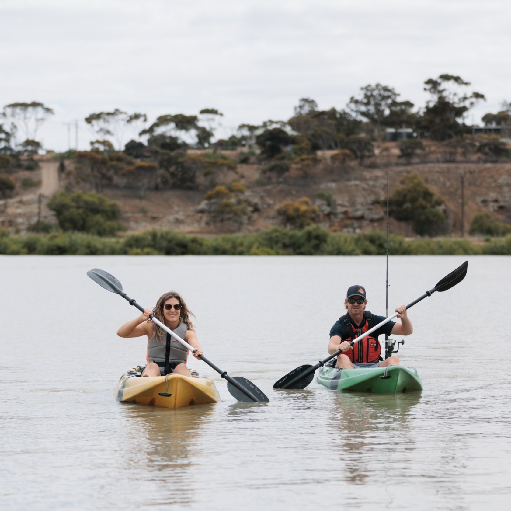 Two people kayaking on a lake with trees in the background.