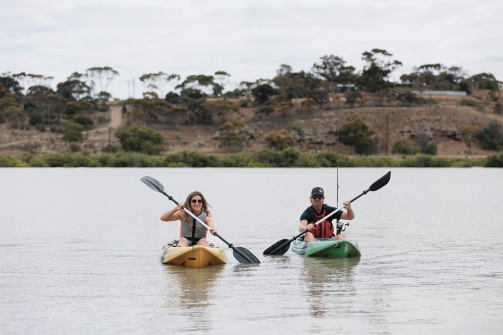 Two people kayaking on a lake with trees in the background.