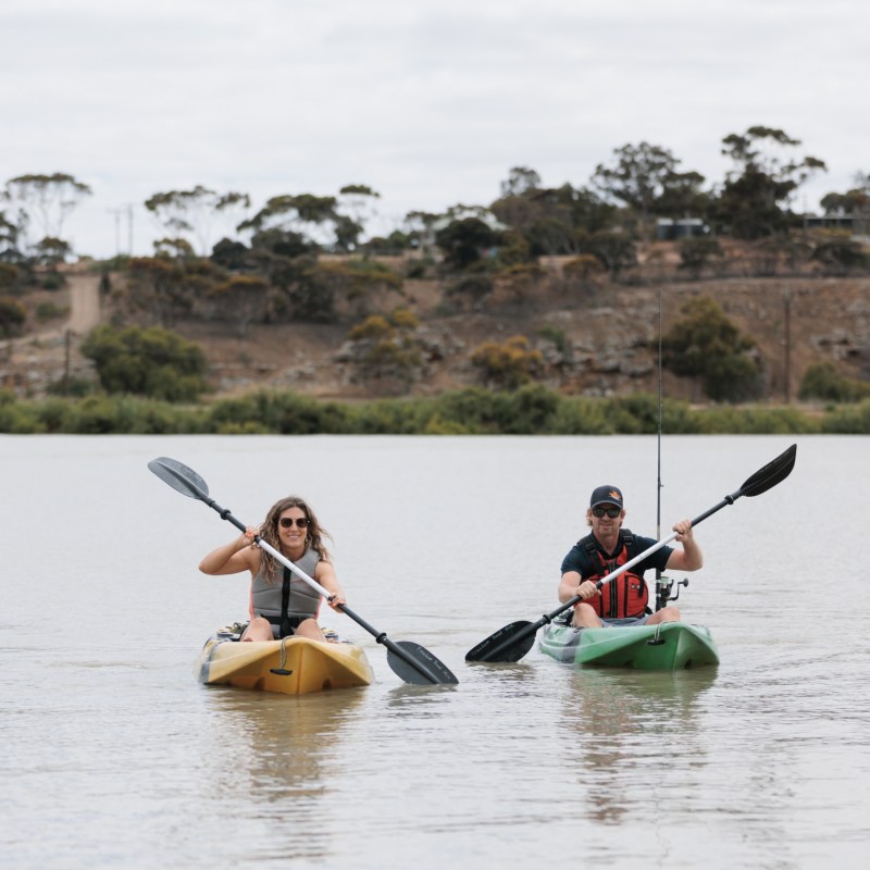 Two people kayaking on a lake with trees in the background.