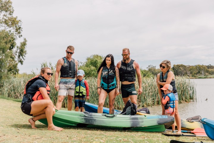 Group of adults and children with kayaks by a lake.
