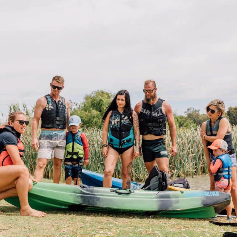 Group of adults and children with kayaks by a lake.