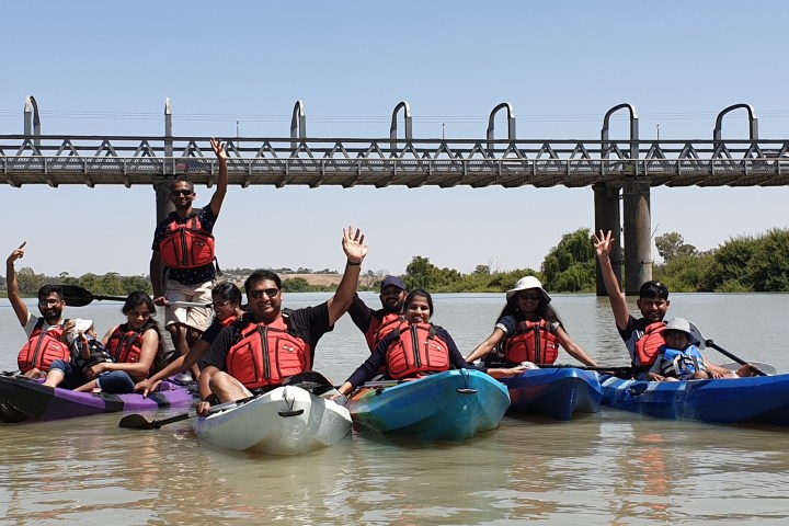 Group kayaking on a river with a bridge in background, people waving.