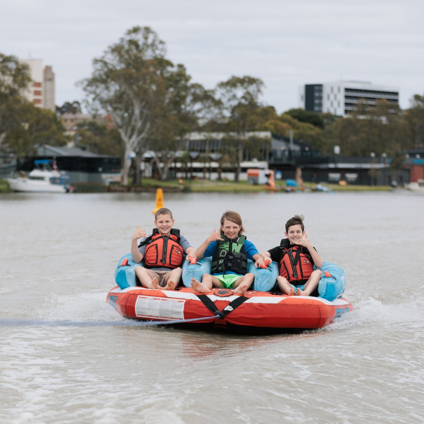Three children ride on an inflatable towable on a river, wearing life jackets, with buildings and trees in the background.