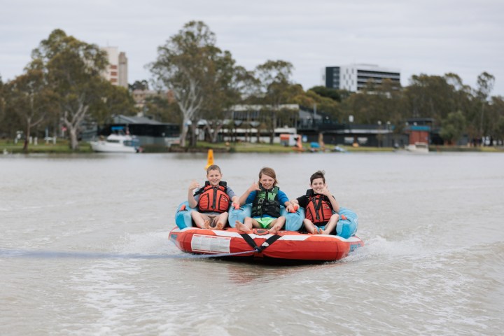 Three children ride on an inflatable towable on a river, wearing life jackets, with buildings and trees in the background.