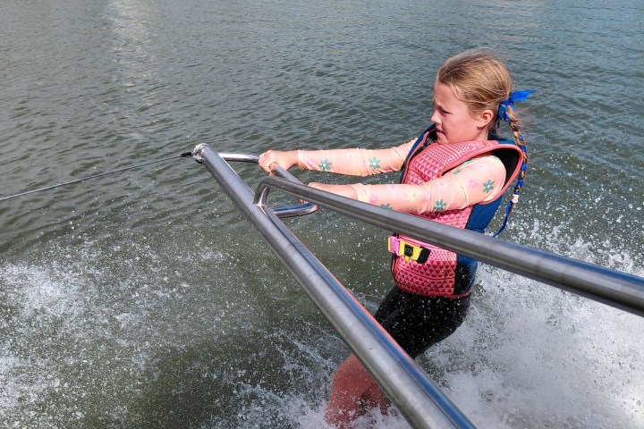 Person water skiing, holding onto a metal bar, with buildings and trees in the background.
