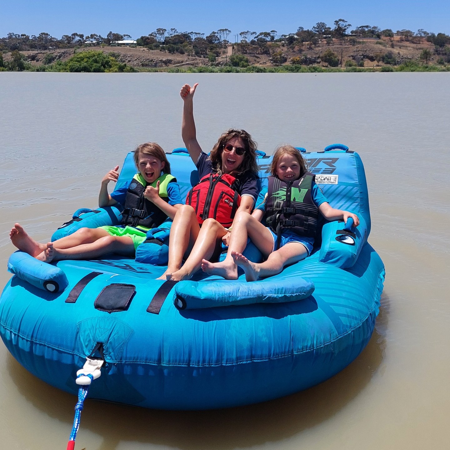 Three people in life vests on a blue inflatable raft on a lake.