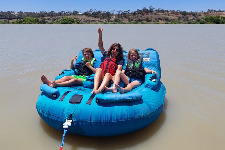 Three people in life vests on a blue inflatable raft on a lake.