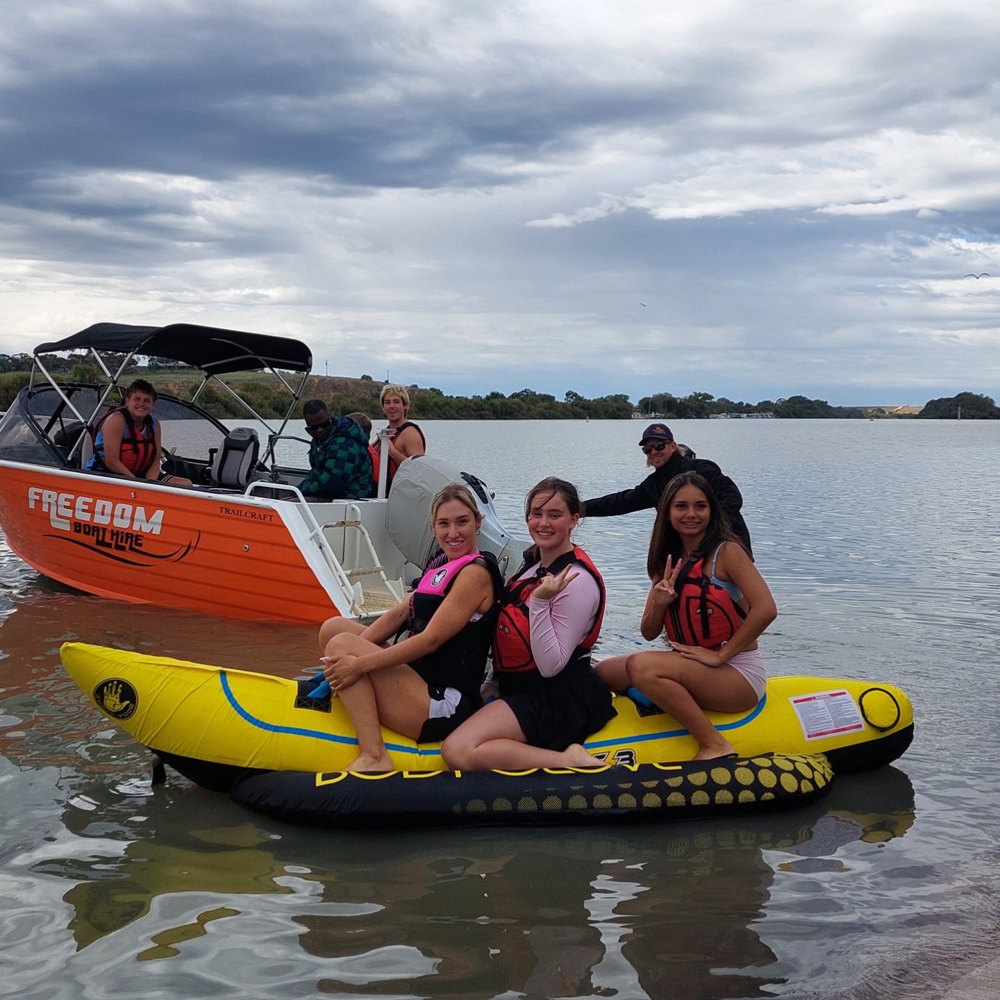 Three people on a banana boat, with a motorboat and two people in the background on a lake.