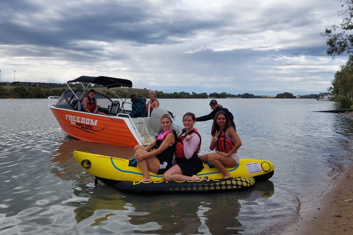 Three people on a banana boat, with a motorboat and two people in the background on a lake.